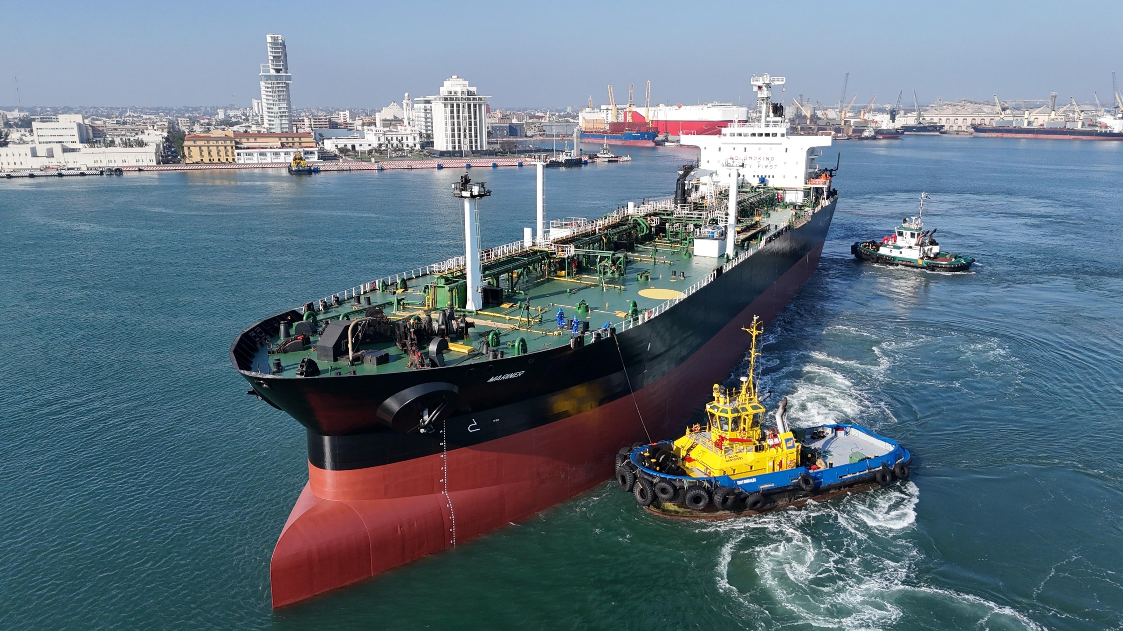 Large oil tanker entering a harbor with tugboats guiding it, city buildings and cranes visible in the background.